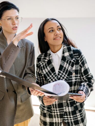 Zwei professionelle Geschäftsfrauen in einem modernen Büro im Gespräch. Eine Frau mit Brille und braunem Karoblazer gestikuliert erklärend, während die andere Frau in schwarz-weißem Mantel Dokumente hält und aufmerksam zuhört.