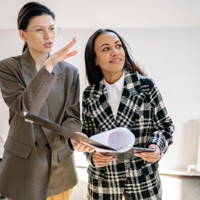 Zwei professionelle Geschäftsfrauen in einem modernen Büro im Gespräch. Eine Frau mit Brille und braunem Karoblazer gestikuliert erklärend, während die andere Frau in schwarz-weißem Mantel Dokumente hält und aufmerksam zuhört.